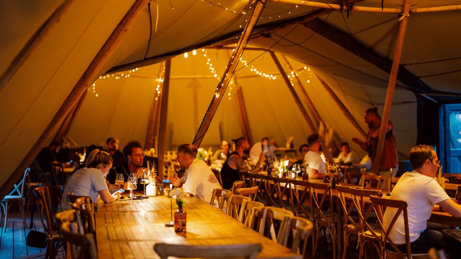 People dining and socializing at long wooden tables inside a warmly lit tipi tent.