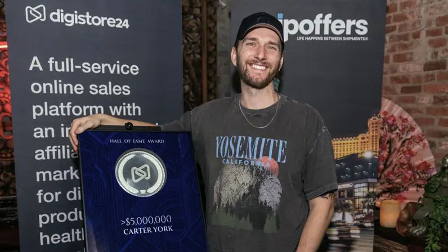 Smiling man Carter York holds a Digistore24 Hall of Fame Award for over $5,000,000 in sales, with company banners behind him.