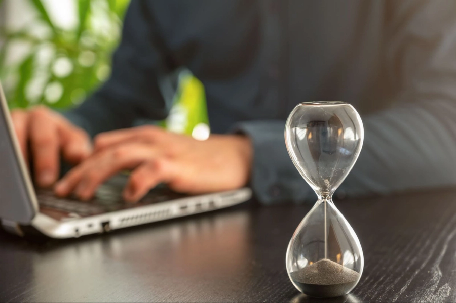 An hourglass with sand nearly run out, and a person typing on a laptop in the blurred background.