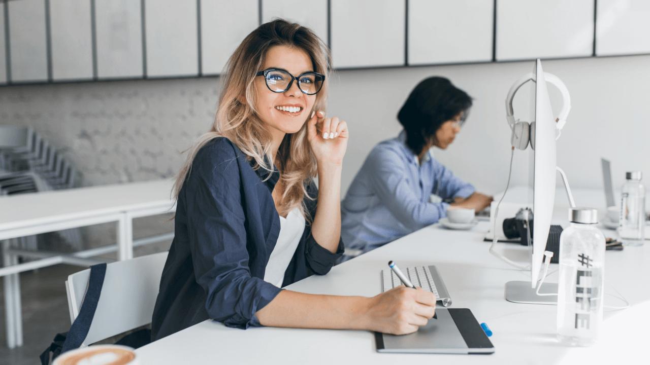 Smiling woman in glasses using a drawing tablet at an office desk.