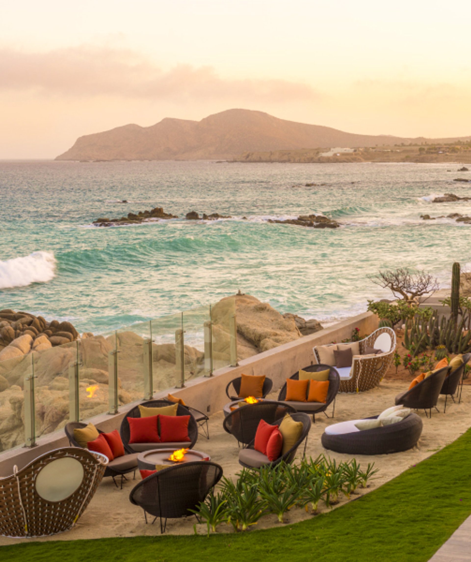 Outdoor lounge with fire pits on a sandy terrace overlooking a turquoise ocean and mountains at sunset.
