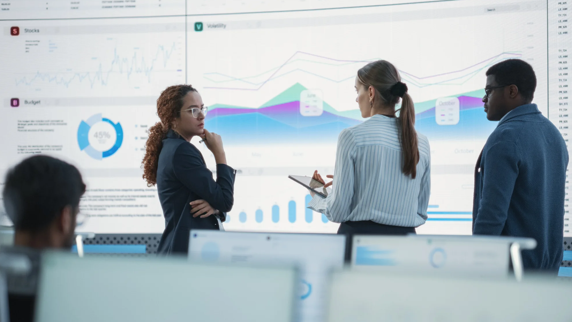 Three business people discuss charts and graphs on a large digital display.