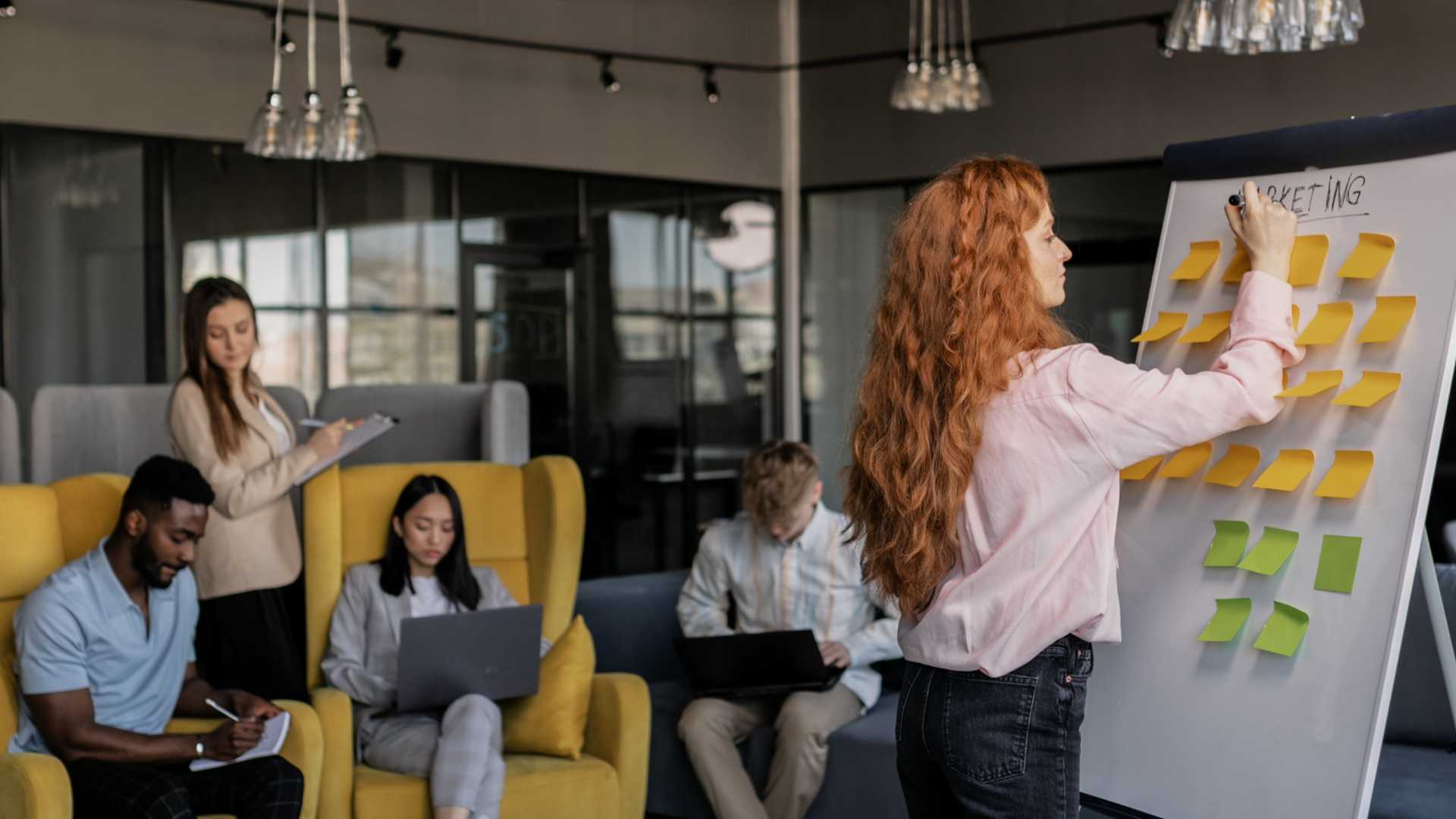 A woman writes on a whiteboard covered in sticky notes while a diverse team works in an office.