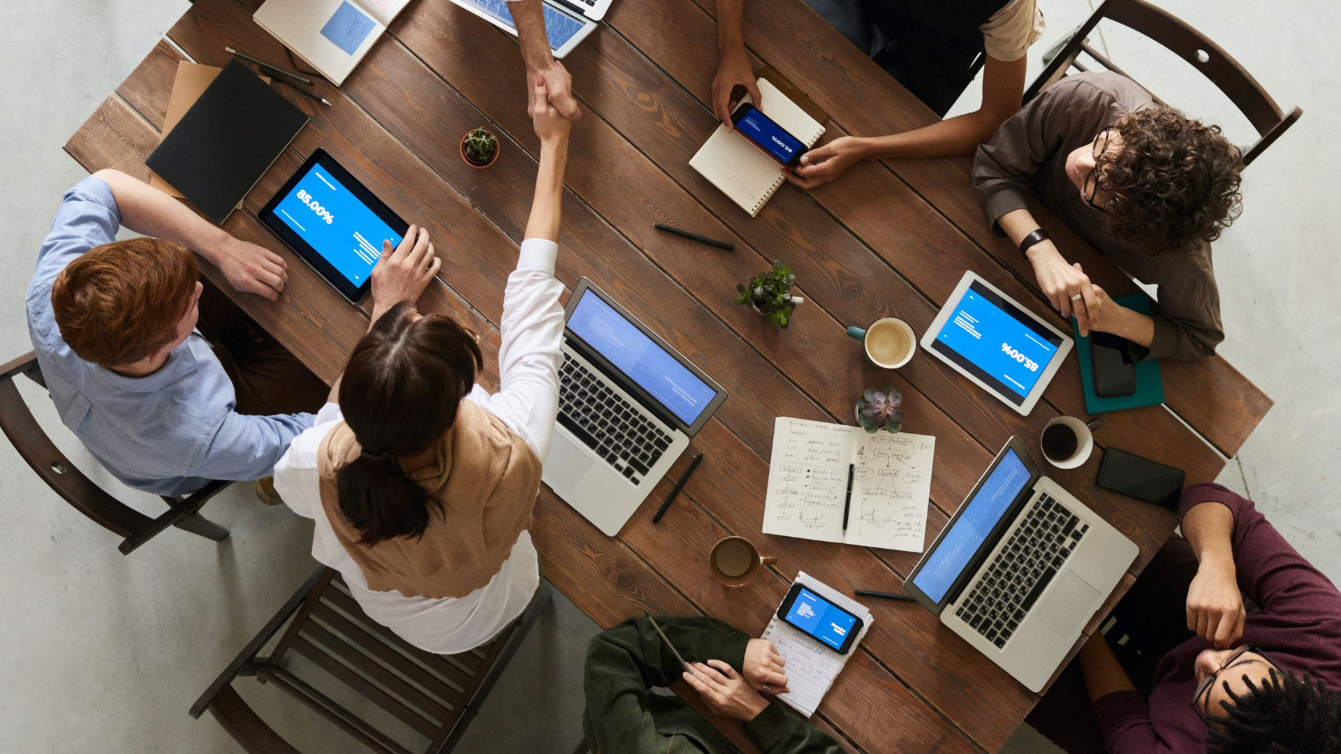 Overhead shot of a group at a table with laptops and tablets; two people shake hands.