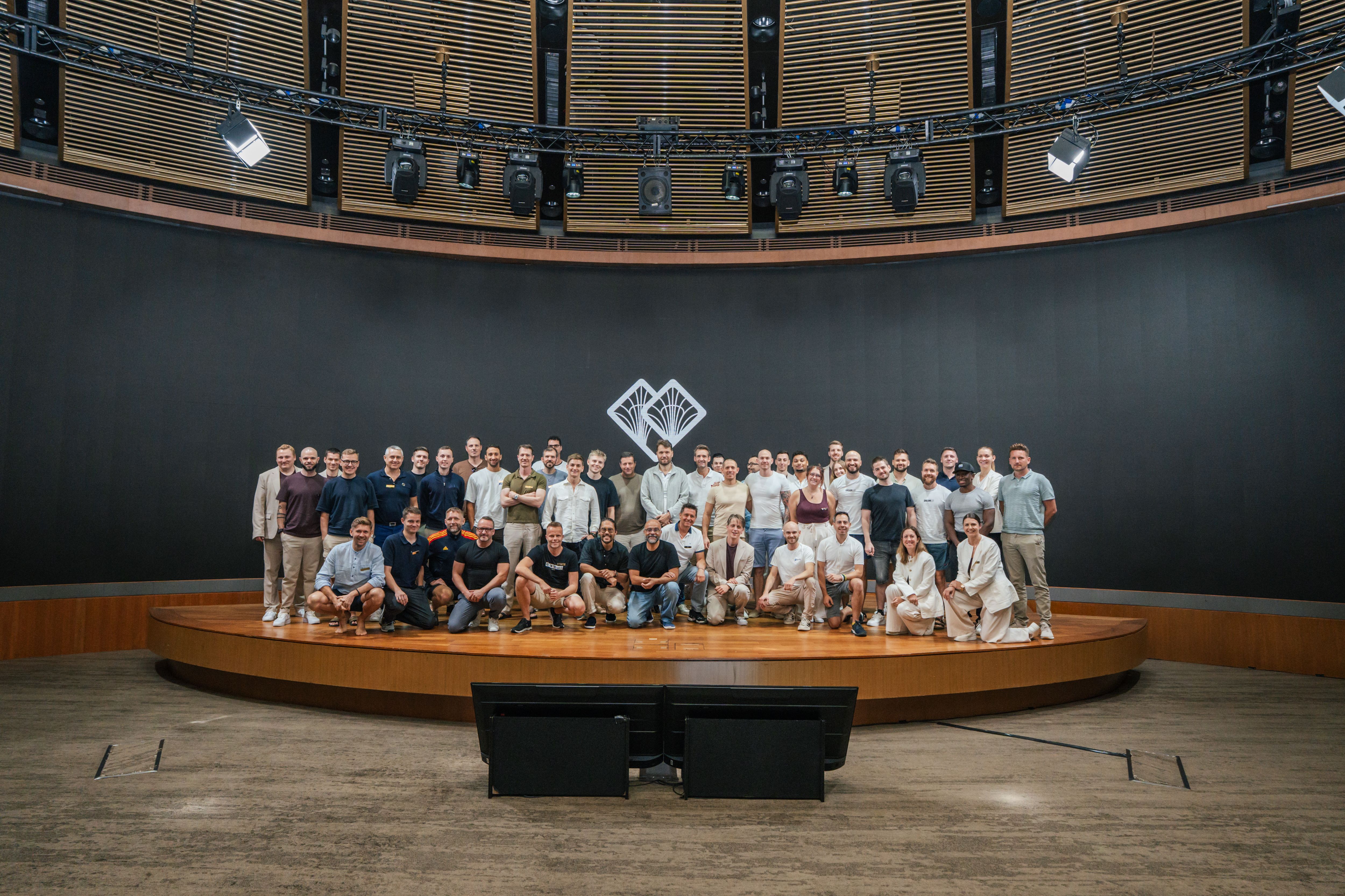 A large group of people, mostly men, pose on a circular stage in a modern auditorium with a white logo on the dark background wall.