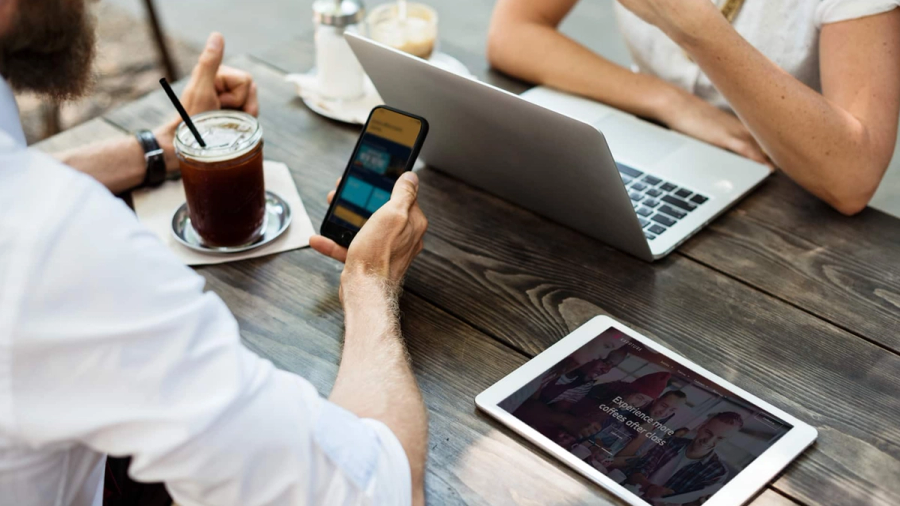 Two people at a wooden table with a laptop, smartphone, and tablet.