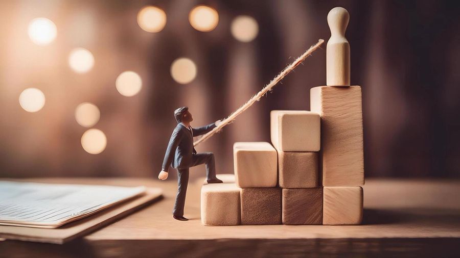 A miniature businessman climbs wooden block steps, holding a rope to a wooden pawn at the top.