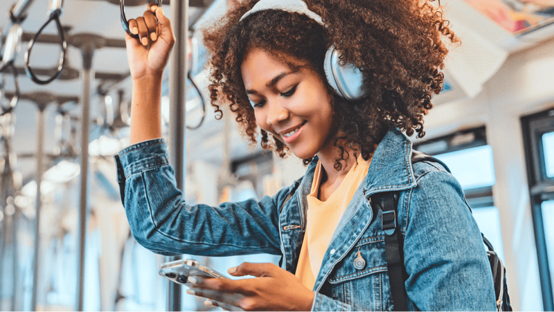 A smiling young woman with headphones and a denim jacket holds a strap and looks at her phone on a train.