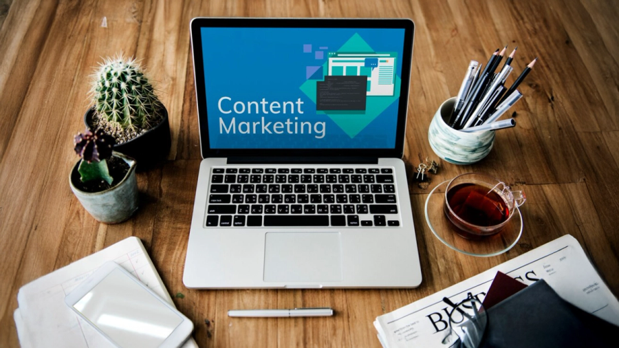Laptop on a wooden desk displays 'Content Marketing,' surrounded by plants, tea, and office supplies.