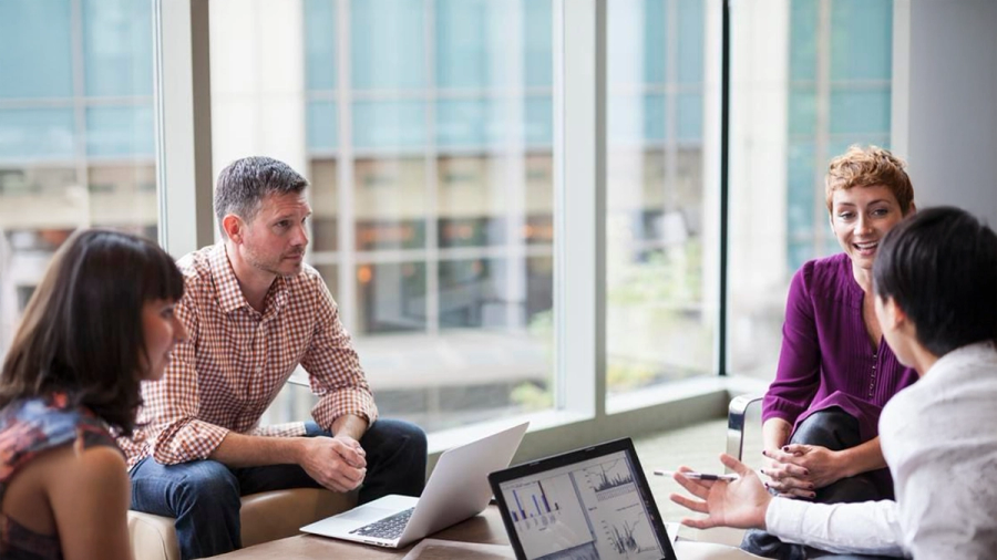 Four colleagues discuss data on laptops in a modern office.