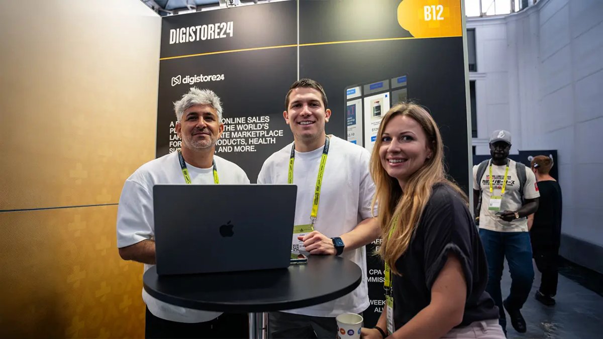Three smiling people at a Digistore24 booth with a laptop.