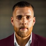 A man with a reddish-brown beard and short haircut looks at the camera, wearing a maroon blazer.