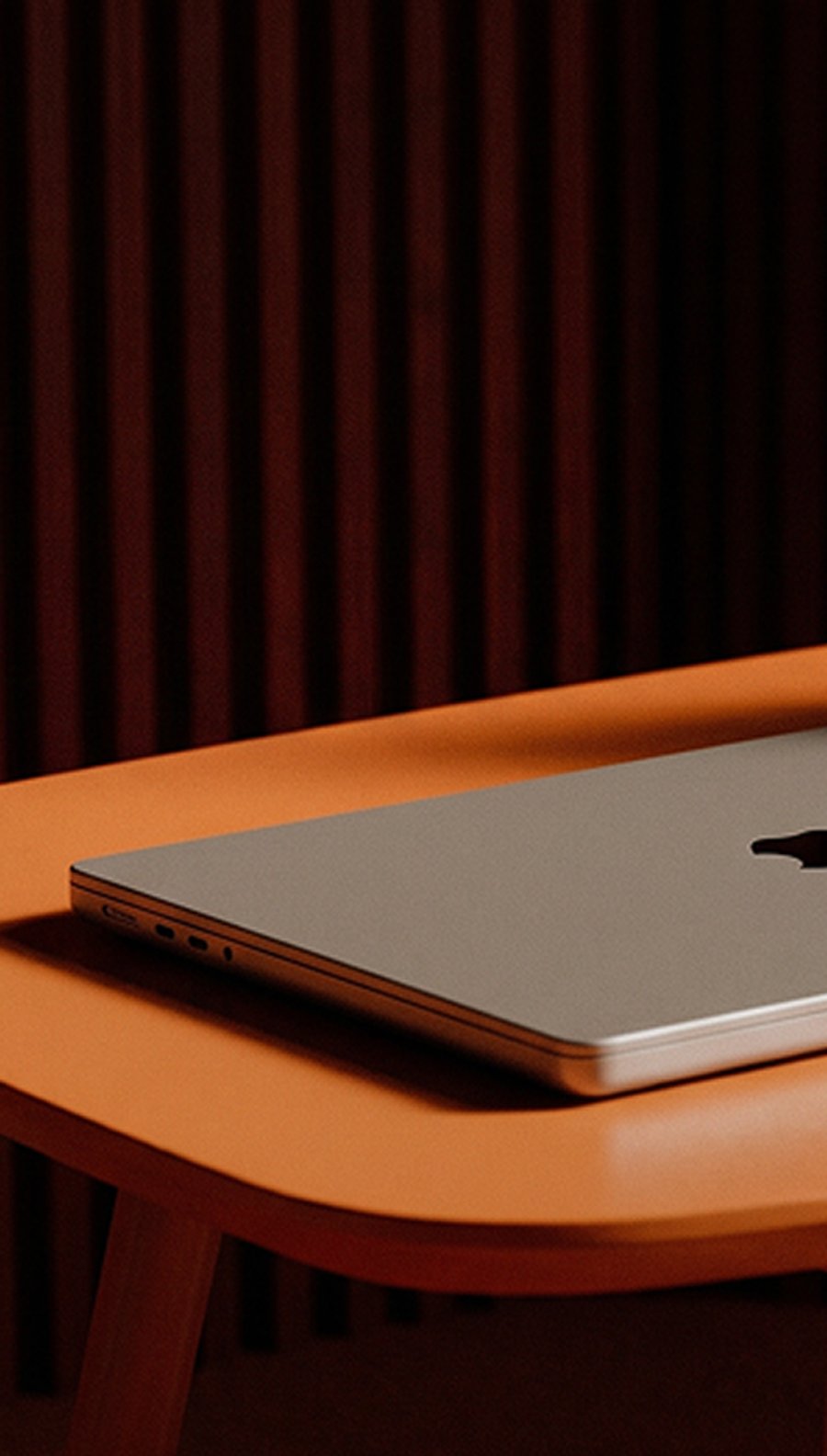 Closed silver MacBook resting on an orange table against a dark, striped background.