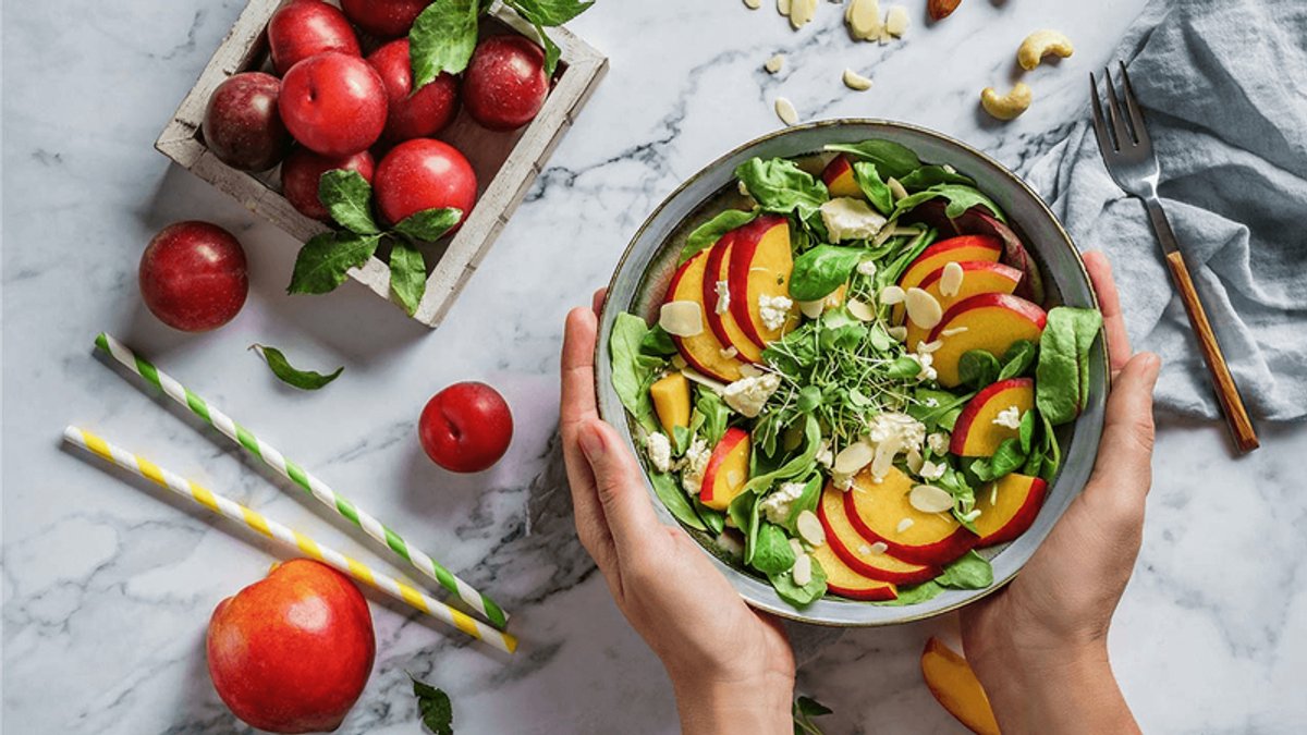 Hands holding a vibrant salad of sliced peaches, greens, and cheese, with plums and nuts on a marble surface.