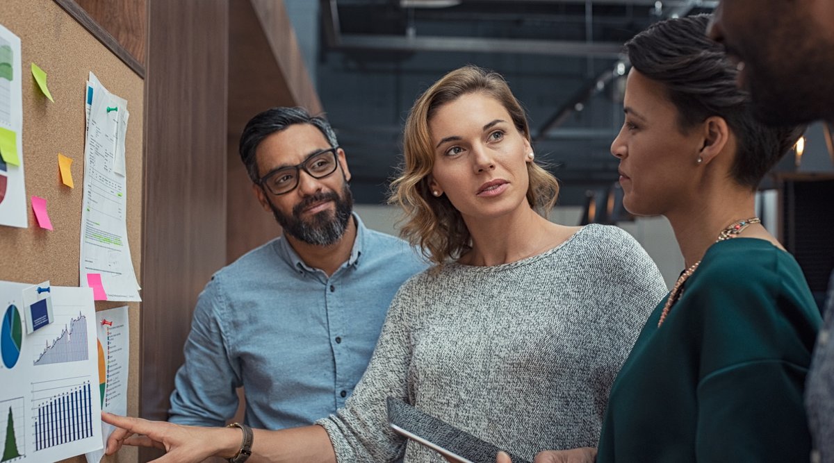 Three colleagues discussing charts on a cork board, with one woman pointing at data.