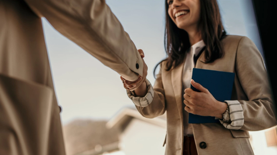 A smiling woman shakes hands with another person, holding a blue book.