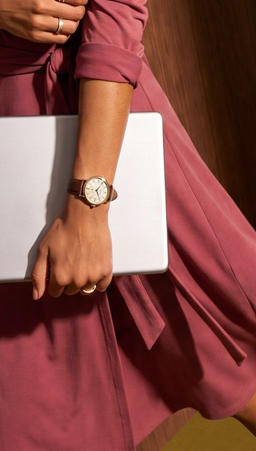 A person in a mauve wrap dress holds a silver laptop, wearing a brown leather watch and gold rings.