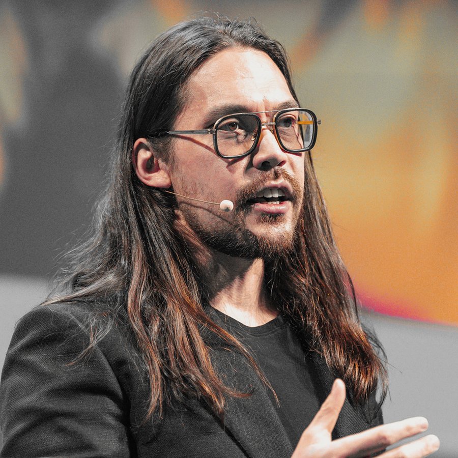 A man with long brown hair, a beard, and glasses wears a black jacket and speaks into a microphone.