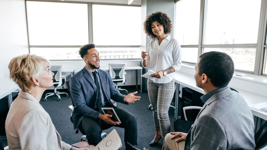 Four professionals in an office meeting, with a woman standing and speaking to three seated colleagues.