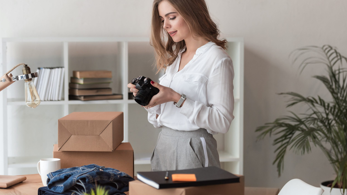 Young woman holding a camera, standing at a desk with shipping boxes and merchandise.