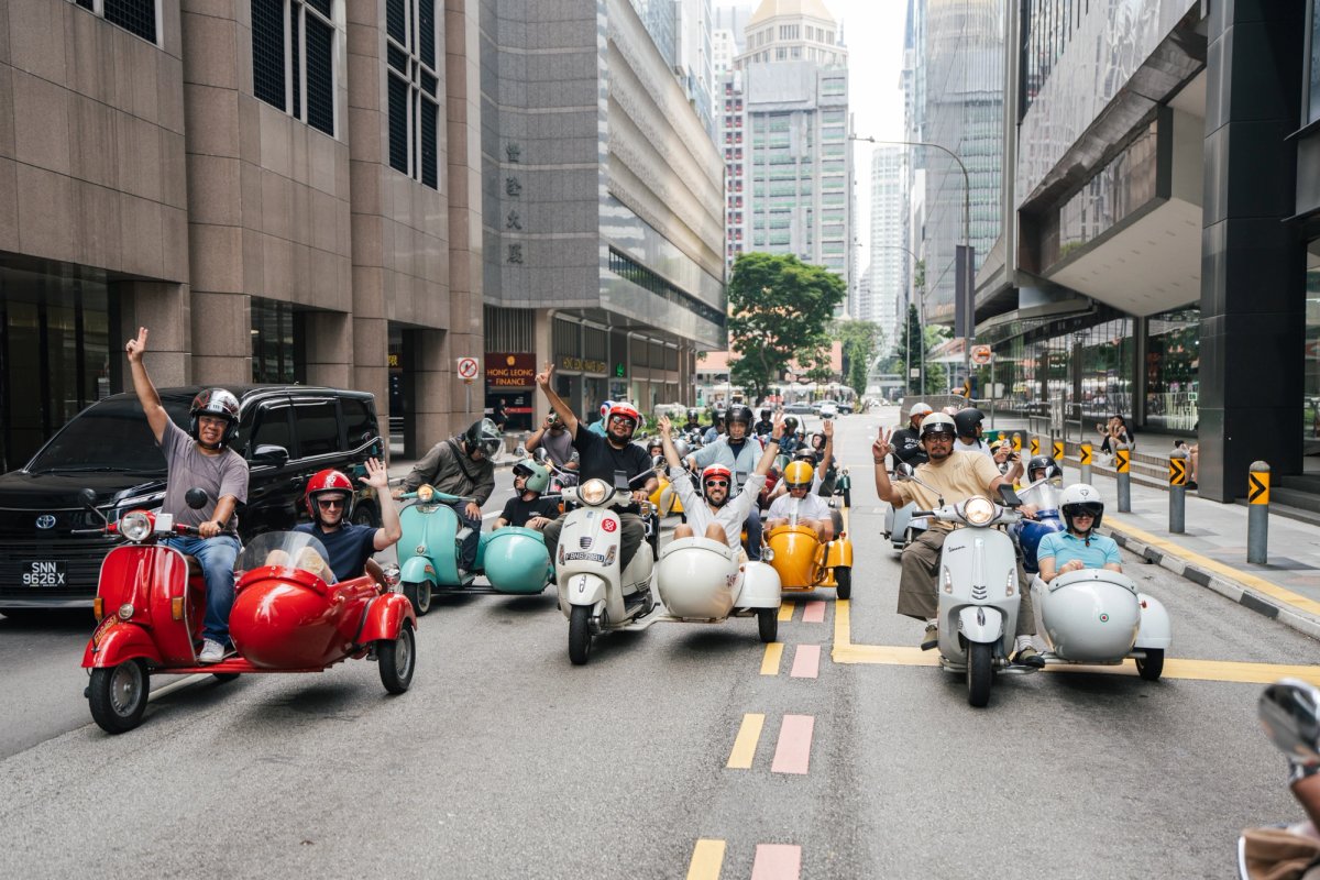 A group rides colorful vintage scooters with sidecars down a city street, waving and smiling.