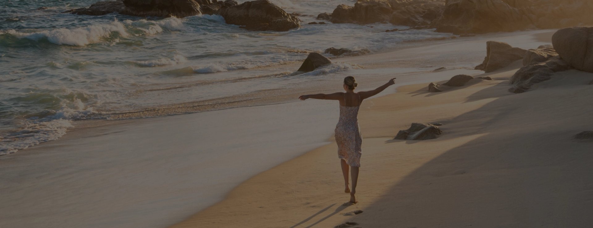 Woman walking on a sandy beach at sunset with arms outstretched, waves crashing nearby.