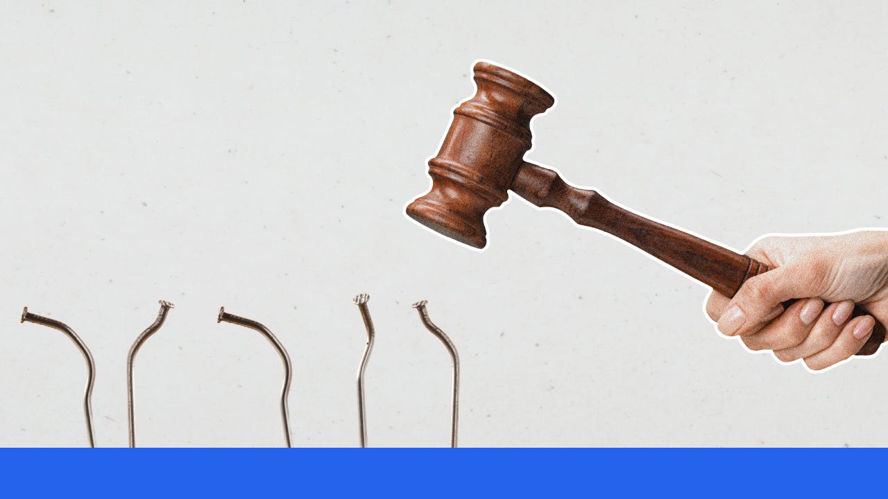 A hand holds a wooden gavel above a row of bent nails.