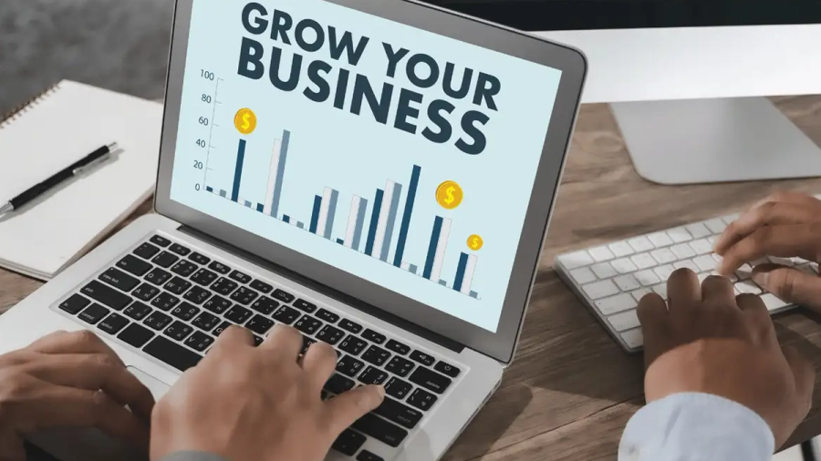 Hands typing on a laptop displaying a "GROW YOUR BUSINESS" chart, with another person typing on a desktop keyboard.