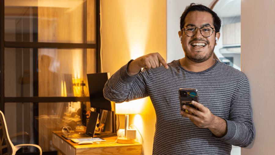 A man wearing glasses and airpods smiles while holding a smartphone and pointing at it.