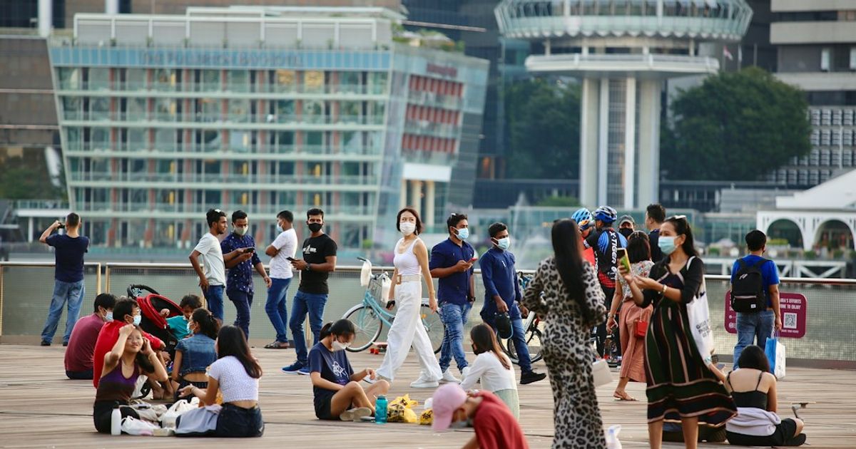 People sit and walk on a wooden waterfront promenade, with glass buildings and a tall tower in the background.