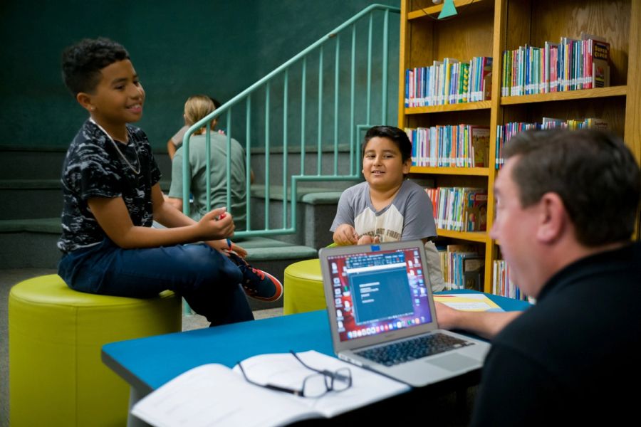 A mentor sits at a table in front of a laptop across from two boys
