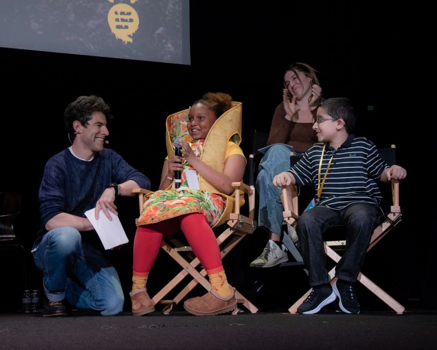 Two students sit in directors chairs, one wears a taco costume and holds a microphone. A man crouches on the floor beside them, smiling