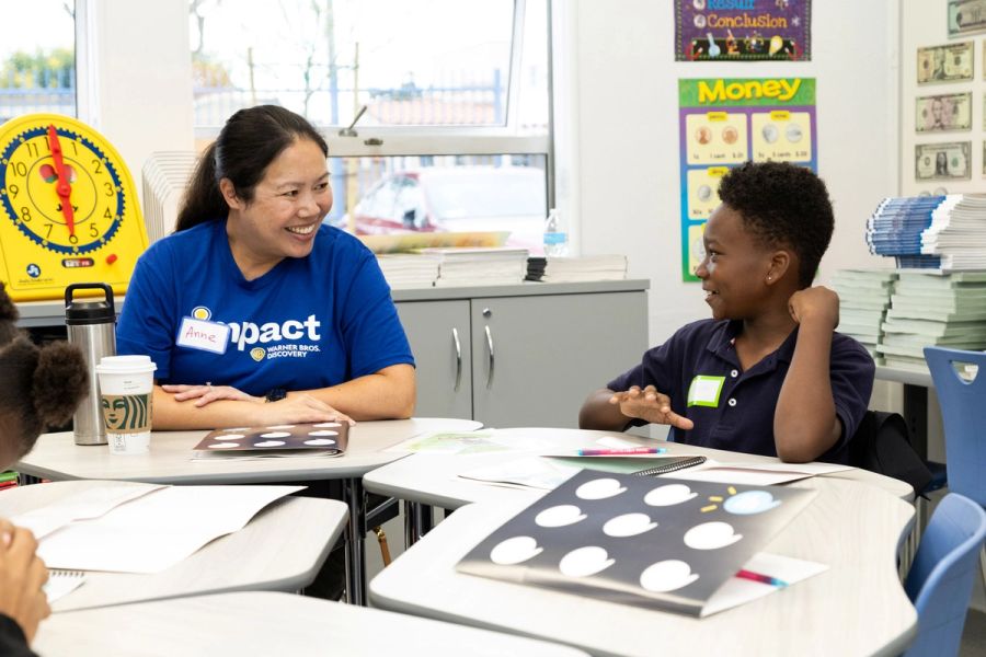 A woman sits next to a boy at classroom desks, both smiling each other