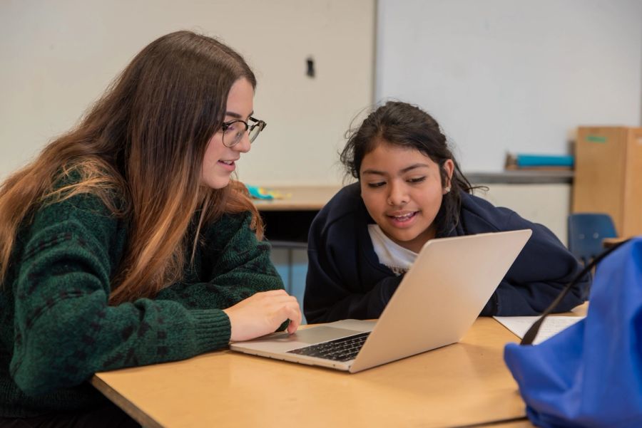 A mentor works at a laptop with a girl, both smiling