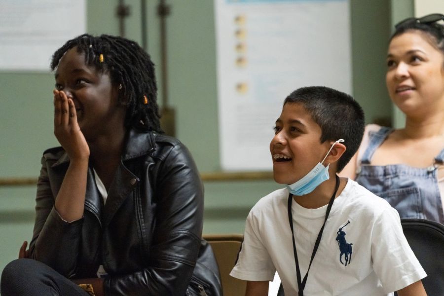 A girl with a hand over her mouth sits next to a boy, both smiling. They look off screen at a stage