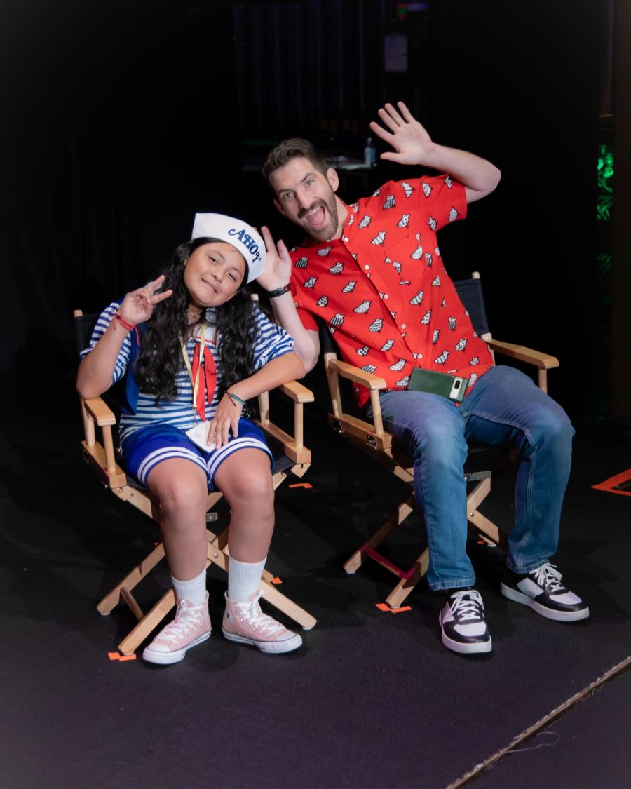 A student and a mentor sit in chairs, smiling and waving at the camera