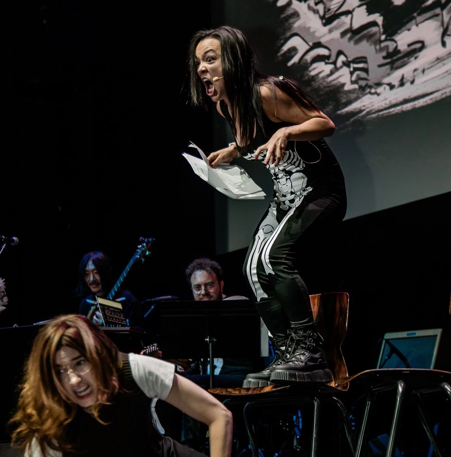 One actor standing on a chair crouches over another actor on the floor