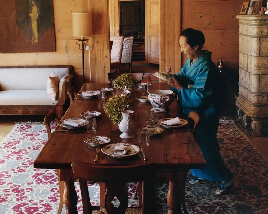 An image from Scenery magazine of Setsuko Klossowska de Rola Photography by Brett Lloyd in her home. She stands beside a vintage wooden dining table set with plates and table decorations