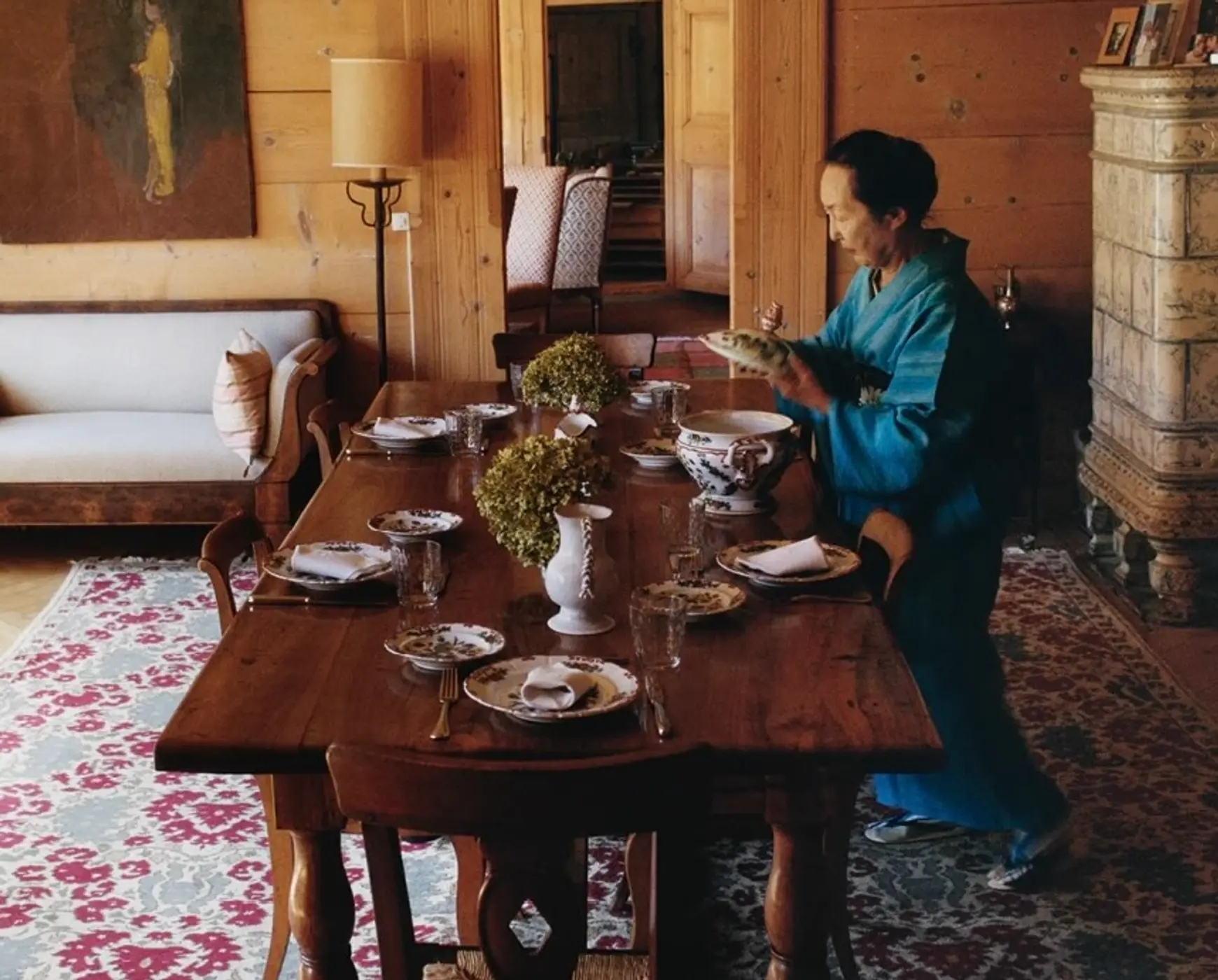 An image from Scenery magazine of Setsuko Klossowska de Rola Photography by Brett Lloyd in her home. She stands beside a vintage wooden dining table set with plates and table decorations