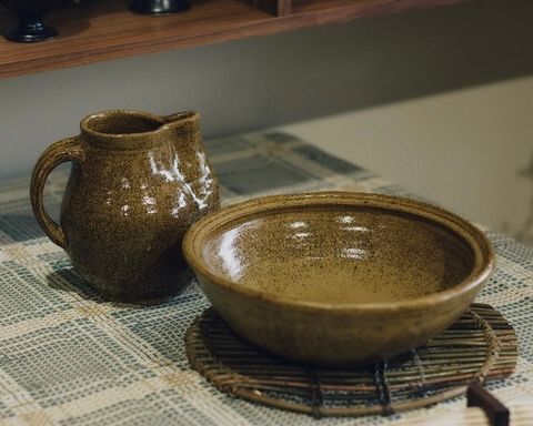 A large ceramic bowl and jug in a green gold speckled glaze created in collaboration between Kirsten Dryburgh and Handcrafted Modern on a dining table with a table cloth onto