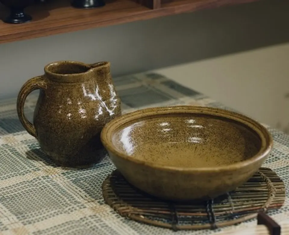 A large ceramic bowl and jug in a green gold speckled glaze created in collaboration between Kirsten Dryburgh and Handcrafted Modern on a dining table with a table cloth onto