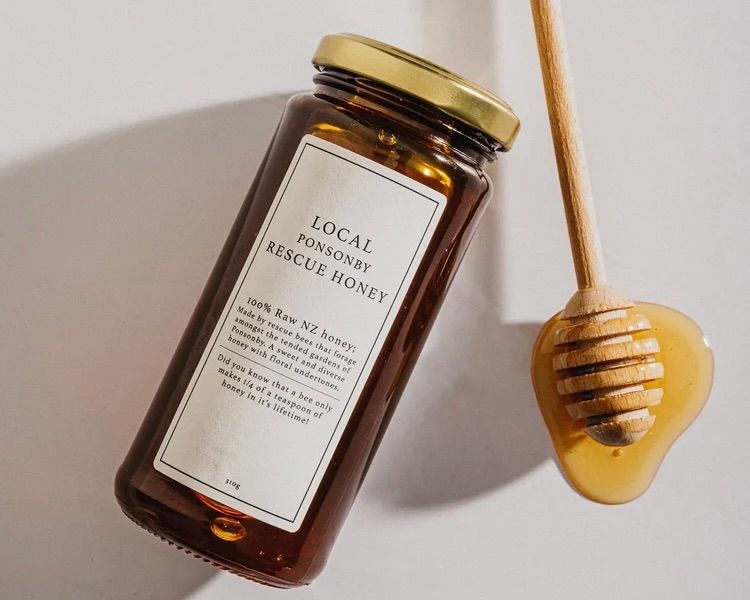 A jar of local Ponsonby New Zealand raw rescue honey in a clear glass jar with a gold lid and wooden honey comb stick next to it from Bees on top.