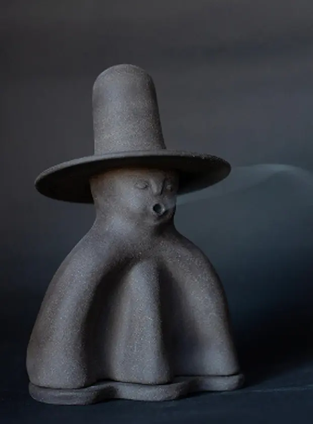 A stoneware ceramic incense holder in the form of a man with a hat with incense smoke coming out of his mouth.