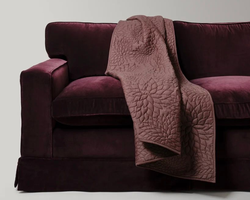 The Dhara Tuberous Throw by Studio Ford, woven from a silk-cotton blend and hand-dyed to a rich chocolate brown, subtle lustre that shifts beautifully with the light draped on a deep purple velvet sofa. 