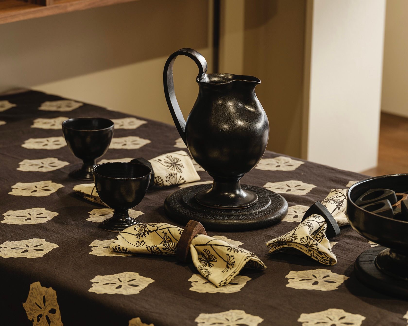 A Blackware jug and two cups on a table setting with a studio ford tablecloth and napkins