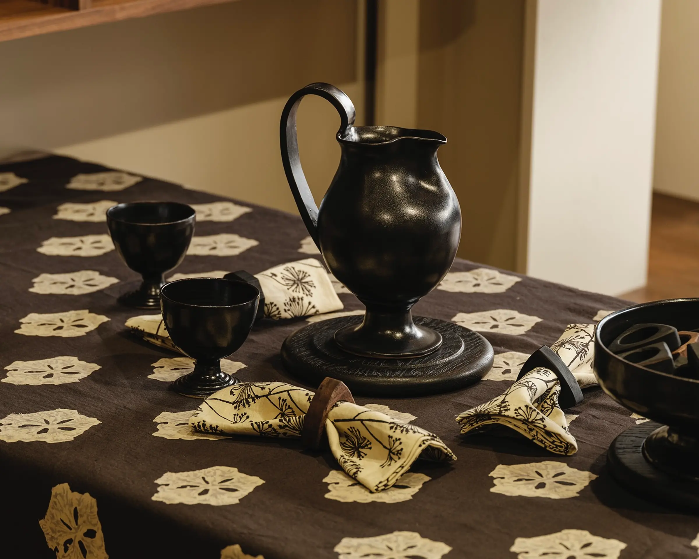 A Blackware jug and two cups on a table setting with a studio ford tablecloth and napkins