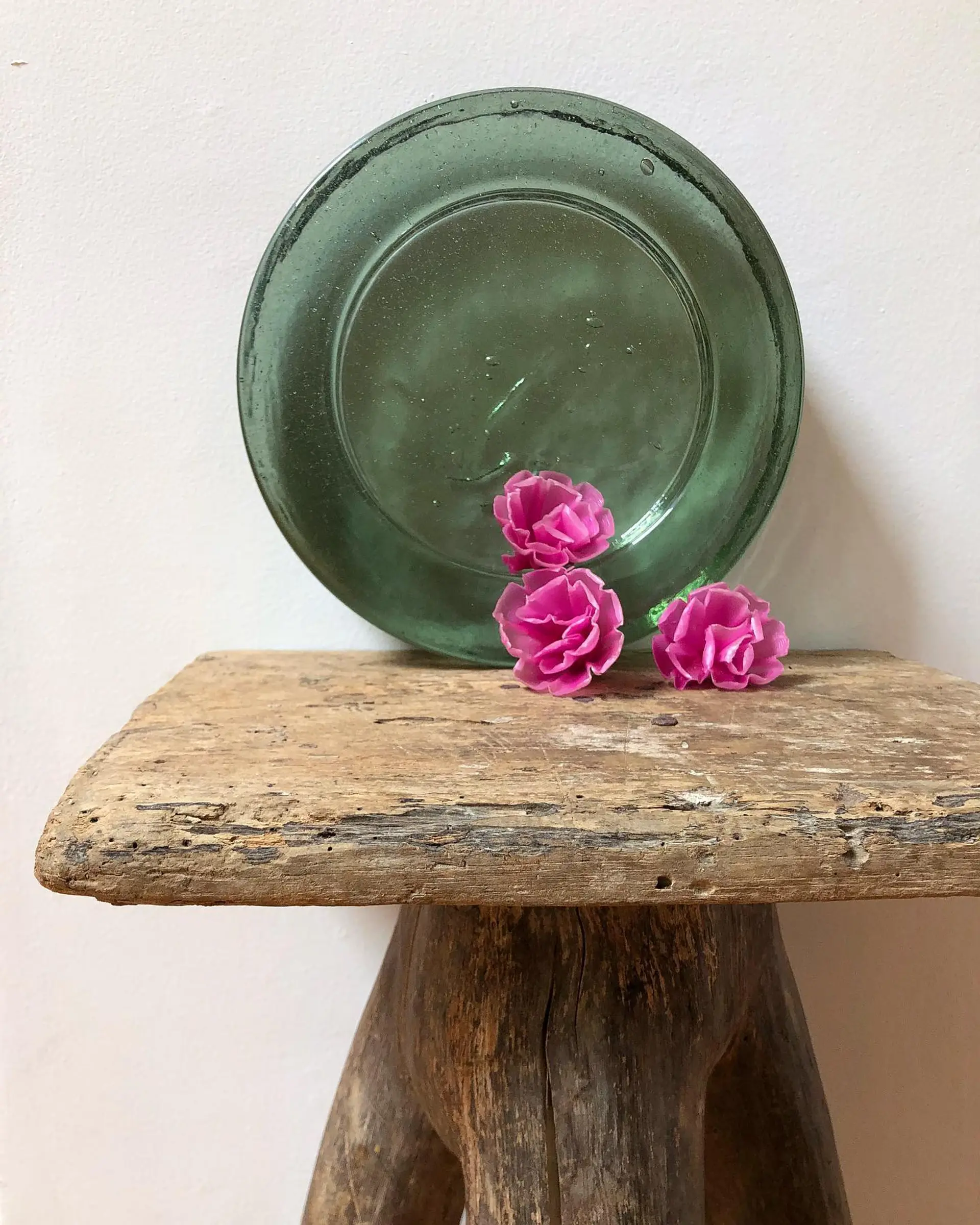 A green catchall glass on a wooden bench with wax flowers infront of it