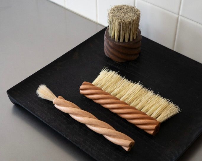 OROS Torsade table brush in turned cherry wood with natural tampico fibres, shown alongside the Torsade kitchen brush and wooden brush on a black timber surface.
