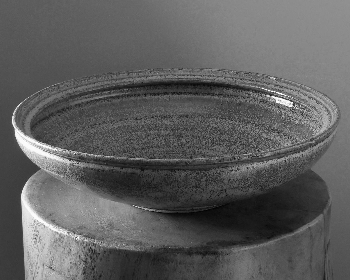A B&W image of a large ceramic bowl in a green gold speckled glaze created in collaboration between Kirsten Dryburgh and Handcrafted Modern on a wooden stool