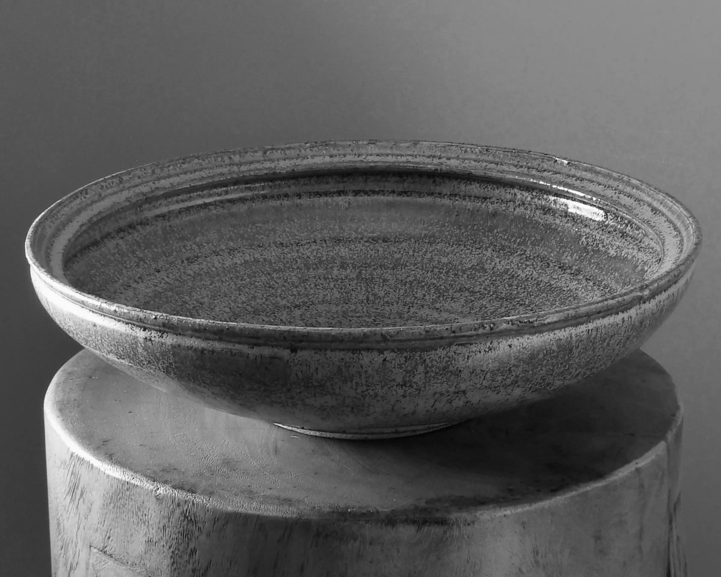 A B&W image of a large ceramic bowl in a green gold speckled glaze created in collaboration between Kirsten Dryburgh and Handcrafted Modern on a wooden stool